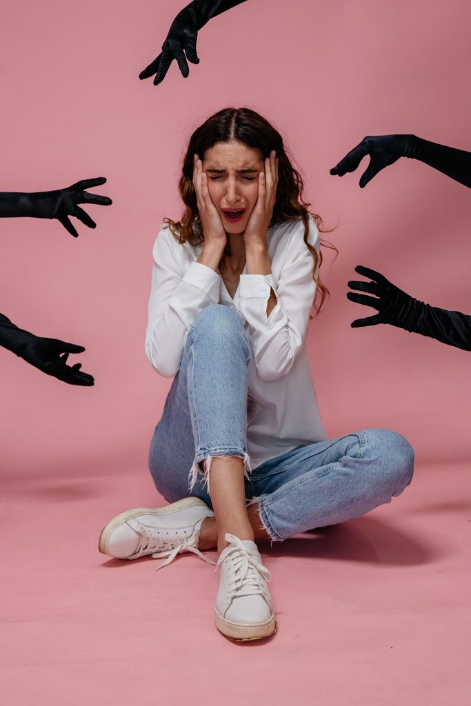 A women sitting down in front of a pink background holding her head between her hands looking stressed
