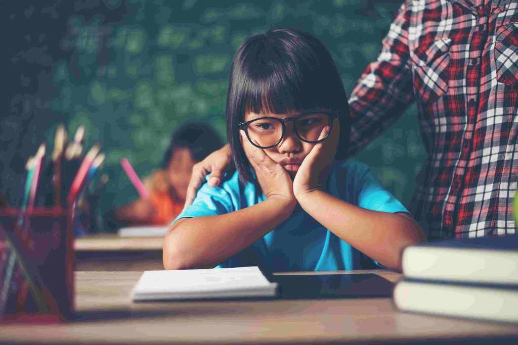 Middle school South East Asian girl with black glasses and a blue t-shirt. Sitting at a desk in school, holding her face in her palms looking discouraged. 